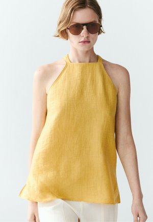Frau mit brauner Sonnenbrille und einem ärmellosen gelben Neckholder-Top mit weißen Hosen vor hellem einfarbigem Hintergrund.