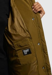 Olijfgroene gewatteerde jas met een glanzende polyester voering, voorzien van een zijzak en een zwart logo label. Drukknoopsluiting is zichtbaar.
