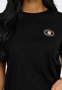 T-shirt noir avec un petit motif d'œil brodé près de l'épaule gauche, porté par une personne aux longs cheveux foncés.