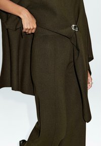 Haut en maille vert foncé avec une main tenant le tissu, doté d'un accent circulaire argenté et d'un pantalon assorti vert foncé. Texture lisse.