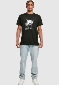 Black t-shirt made of cotton featuring a wolf graphic in gray and white on the front. The model wears light blue jeans and white sneakers.