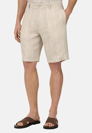 Shorts de lino beige con finas rayas verticales blancas, usados por una persona de pie con las manos en los bolsillos y sandalias marrones en los pies.