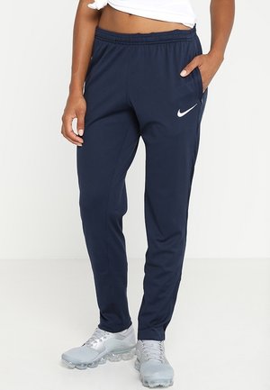 Person trägt dunkelblaue Nike-Sweathose und hellgraue Nike-Sneakers, steht mit einer Hand in der Tasche vor einfarbigem Hintergrund.