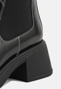 Gros plan sur une bottine noire à talon bloc épais, panneau latéral élastique et surpiqûres blanches sur cuir lisse.