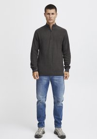 Pull zippé gris foncé avec texture côtelée, manches longues et col montant, associé à un jean bleu clair et des chaussures grises.