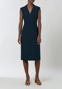 Femme portant une robe bleu marine sans manches, longueur au genou, avec un décolleté en V, et des sandales marron à talons blocs ouverts, debout devant un fond uni.