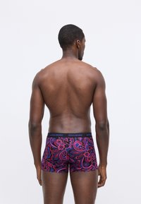 Muž s tmavou pokožkou otočený chrbtom, oblečený v farebne vzorovaných boxerách s ružovými, fialovými a čiernymi kvetinovými vzormi na bielom pozadí.