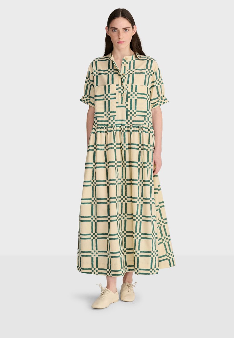 Femme portant une longue robe à carreaux crème et verte avec manches courtes et des baskets crème, debout devant un fond clair uni.