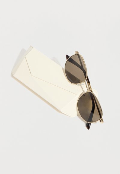 Γυαλιά ηλίου αεροπόρου με χρυσό πλαίσιο και σκούρους φακούς, τοποθετημένα διαγώνια πάνω σε λευκή διπλωμένη θήκη για γυαλιά.