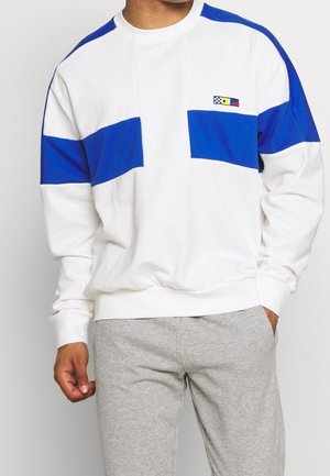 Mężczyzna ubrany w biały sweter z niebieskimi poziomymi paskami i małym wielokolorowym logo na piersi, w połączeniu z jasnoszarymi spodniami dresowymi.