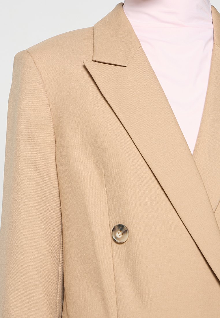 Beige Doppelreiher-Blazer mit Reverskragen, der eine glatte Textur und ein klares Knopf-Detail in der Taille aufweist.
