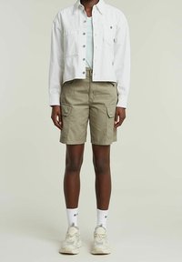 Veste en denim blanc, chemise vert clair, short cargo kaki avec poches latérales, chaussettes blanches montantes et baskets beiges, posant devant un fond neutre.