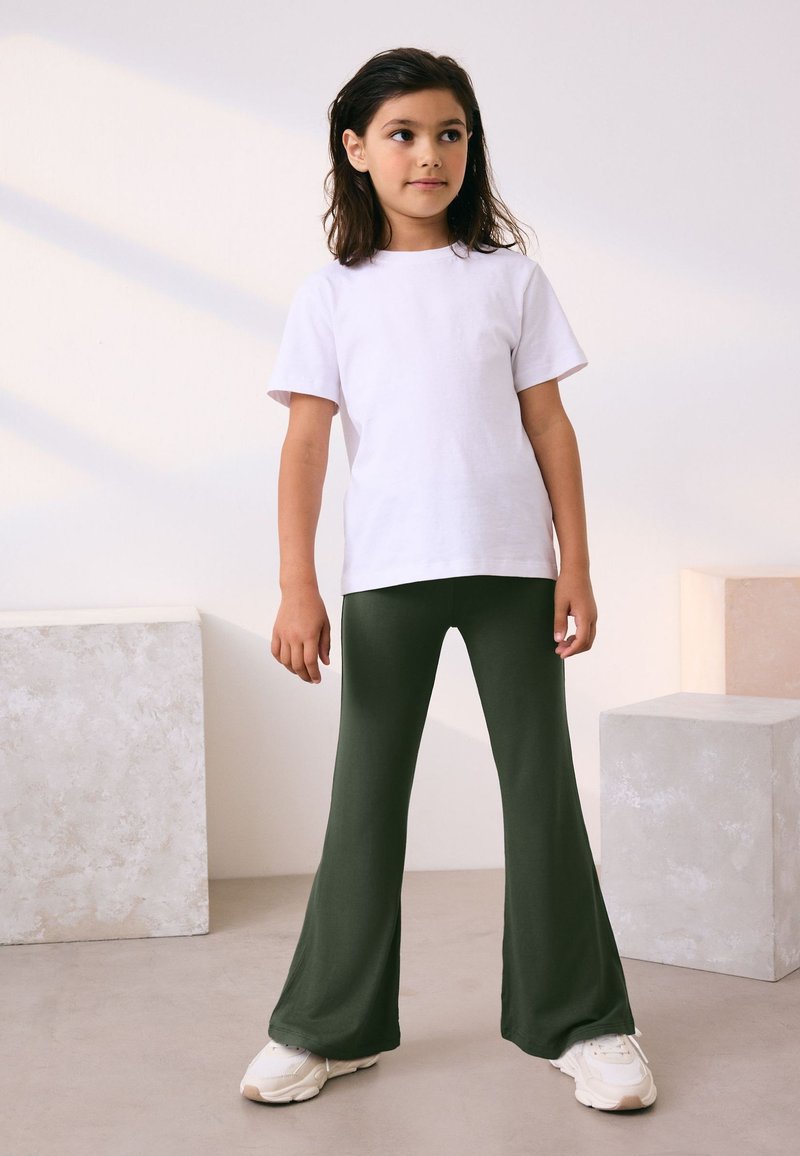 Fiatal lány belül áll, fehér rövid ujjú pólót, sötétzöld bőrszárú nadrágot és fehér sportcipőt visel, kissé oldalra néz.