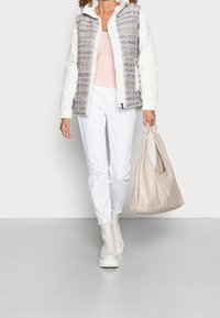 Chaqueta acolchada blanca tipo puffer con acentos de tweed multicolor, blusa rosa claro, pantalones blancos ajustados y bolso ancho beige. Botas blancas de suela gruesa.
