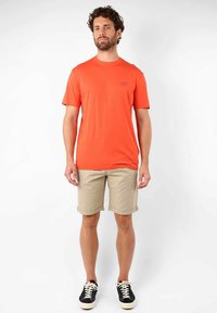 T-shirt orange à manches courtes avec un col rond, associé à un short beige et des baskets noires. Présente un petit logo sur le t-shirt.