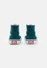 Tummansininen mokkanahkainen korkeavartinen lenkkari, jossa on valkoisia yksityiskohtia ja vetskari takana; varustettu kumipohjalla, jossa on punainen "Vans Off The Wall" -logo.