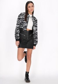 Chaqueta bomber de patrón de cebra en blanco y negro, corte recortado, superpuesta sobre una camisa blanca, combinada con una falda mini de cuero negra y botas con cordones.