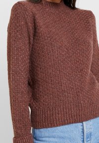 Gros plan d'un pull en maille marron texturée avec des poignets et une base côtelés, porté avec un jean en denim bleu clair.