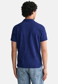 Navy blue short-sleeve polo shirt with a classic collar, made of a smooth fabric. Back view shows fitted design and subtle side slits.