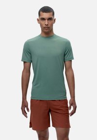 Īsām piedurknēm, apaļas kakla formas krekls no norāmīgas zaļas auduma, kombinēts ar oranžiem šortiem. Materiāls šķiet viegls ar gludu tekstūru.