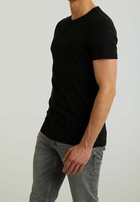 Homme portant un t-shirt noir uni à manches courtes et un jean gris, debout devant un fond gris clair avec une main détendue le long du corps.