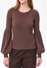 Vrouw draagt een nauwsluitende bruine geribbelde trui met ballonmouwen en bijpassende bruine broek tegen een effen witte achtergrond.