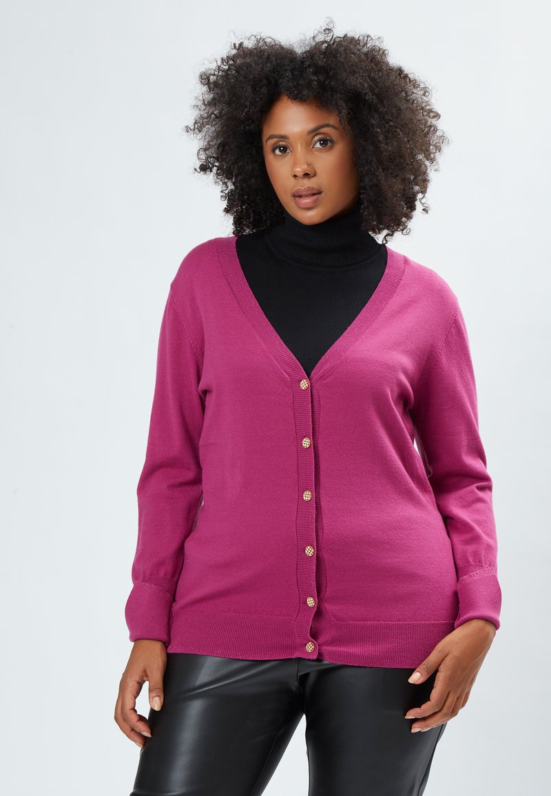 Cardigan fuchsia en tissu doux, avec un col en V, des manches longues et des boutons dorés sur le devant. Porté sur un turtleneck noir.