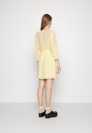 Femme aux longs cheveux bruns portant une robe jaune clair, des chaussettes vertes à motif cœur, des chaussures à plateforme noires et des boucles d’oreilles vertes, debout contre un mur blanc.