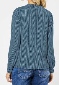 Blå långärmad topp med ett chevronmönster i mörkare blått. Har en rund halsringning och pucklade ärmar. Parat med blå denimjeans.