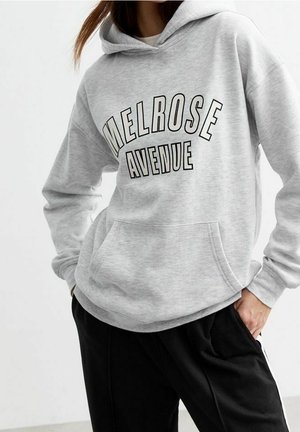 Osoba ubrana w jasnoszarą bluzę z kapturem z napisem "MELROSE AVENUE" i czarne spodnie, pozująca z rękami w kieszeniach na jednolitym tle.