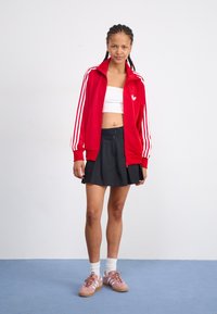 Rote zip-up Trainingsjacke mit weißen Streifen, weißem Logo, getragen über einem weißen Crop-Top. Schwarzer plissierter Rock und rosa Sneakers mit weißen Akzenten.