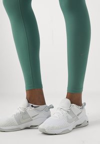 Zelene tajice s glatkom teksturom i kontrastnim šavovima, uparene s bijelim Nike tenisicama koje imaju teksturiran gornji dio i sive detalje.