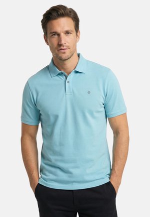 Man met lichtbruin haar, gekleed in een lichtblauw poloshirt met korte mouwen en zwarte broek, staand met zijn handen in zijn zakken.