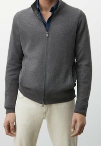 Graue Strickjacke mit Reißverschluss und strukturiertem Muster, gerippte Bündchen und Saum, getragen über einem blauen Hemd, kombiniert mit hellen Hosen.