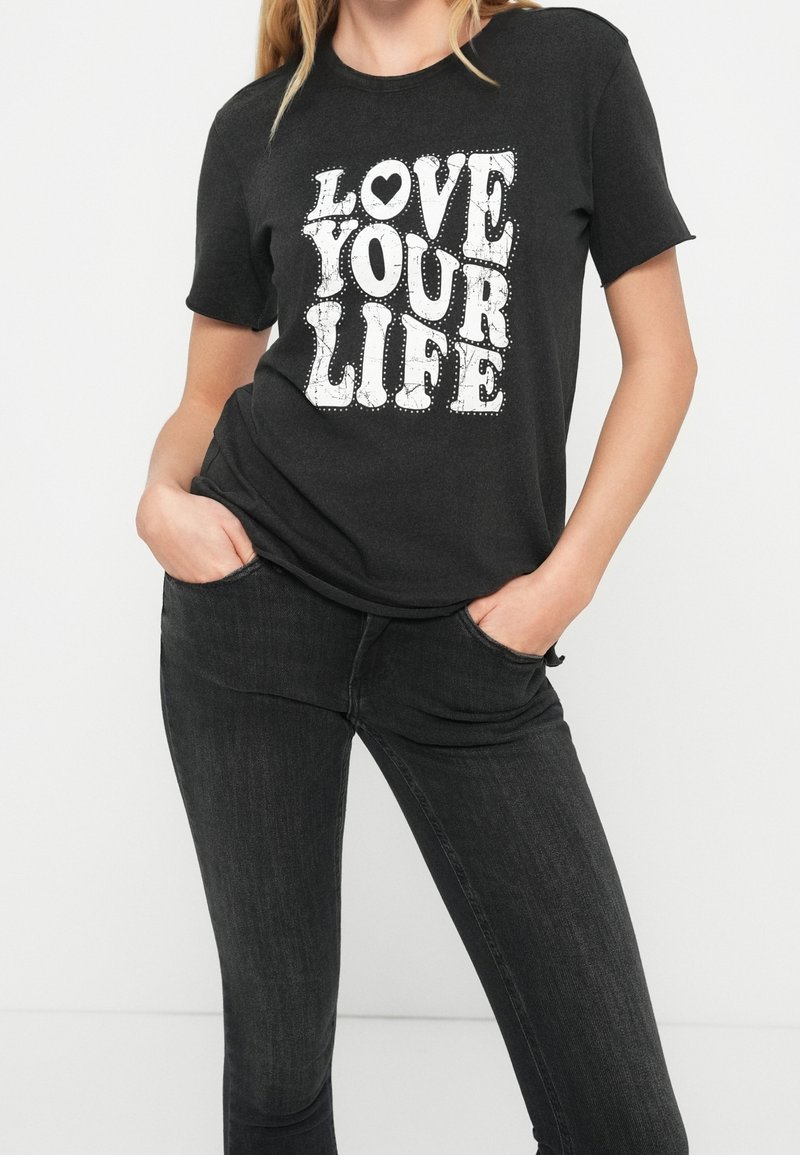 Femme portant un t-shirt noir avec le texte blanc "LOVE YOUR LIFE" et un jean noir, mains reposant dans les poches avant.