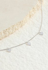 Náhrdelník so strieborným príveskom v tvare srdca na retiazke, ktorý obsahuje tri malé srdcové prívesky s hladkým, lesklým povrchom. Nasnímané na textúrovanom pozadí.