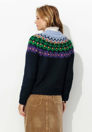 Pull en tricot avec un motif de neige coloré en vert, violet et orange sur un corps bleu marine, avec un col rond et des manches longues.