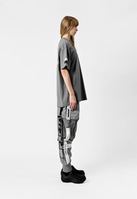 Camiseta gris de gran tamaño con mangas cortas, combinada con pantalones cargo grises que cuentan con múltiples bolsillos y etiquetas en blanco y negro contrastantes. Zuecos negros.