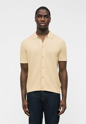 Homme portant une chemise en maille beige à manches courtes avec des boutons et un jean bleu foncé, debout devant un fond clair uni.