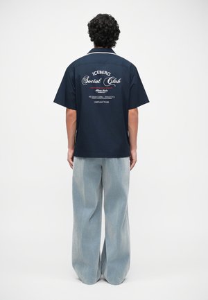 Navy blue short-sleeved shirt with white trim and printed text on the back; paired with wide light blue denim pants.