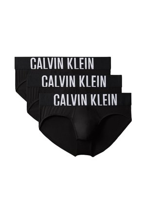 Gespiervde man staat blootsvoets, draagt zwarte Calvin Klein slips tegen een effen grijze achtergrond.