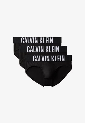 Gespiervde man staat blootsvoets, draagt zwarte Calvin Klein slips tegen een effen grijze achtergrond.