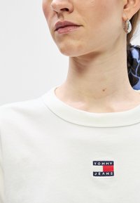 Biela mikina s okrúhlym výstrihom, ktorá obsahuje malé vyšívané logo "TOMMY JEANS" v námorníckej modrej, červenej a bielej farbe. Hladká textúra, neformálny dizajn.