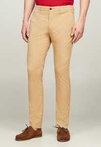 Chinos beige con una vestibilità slim, realizzati in cotone. Presentano una parte frontale piatta, chiusura con bottone e tasche laterali. Indossati con scarpe mocassino marroni.