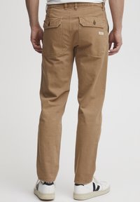 Lichtbruine katoenen broek met rechte pijpen, achterzakken, versterkte naden en subtiele branding. Gedragen met witte sneakers.
