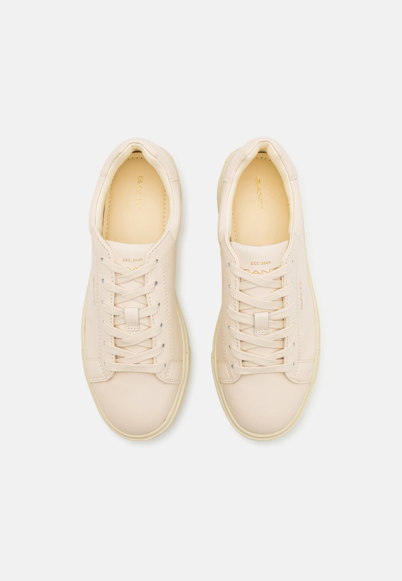Zapatillas de cuero color beige claro con una textura suave, puntera redondeada, seis ojetes y cordones en tono similar. Logo dorado en relieve en la lengüeta.