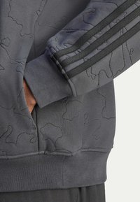 Harmaa collegepaita, jossa on kohokuvioitu abstrakti kuvio, resorit hihansuissa ja sivutaskut. Mustat raidalliset yksityiskohdat hihoissa lisäävät tyyliä.