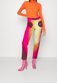 Farbverlaufshosen, die von Pink nach Gelb gehen, mit großen braunen Rundtaschen. Kombiniert mit einem langen orangen Oberteil und rosa High Heels.