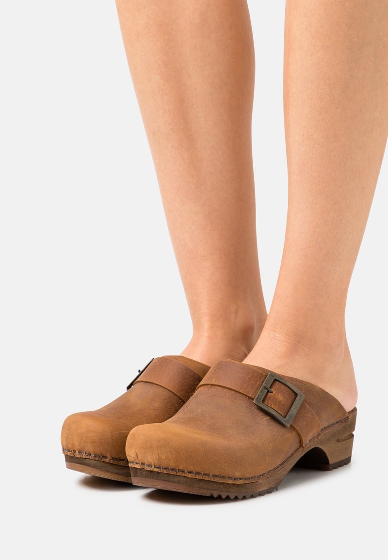 Personne portant des sabots en cuir marron avec bride à boucle et petit talon, debout sur un fond clair.