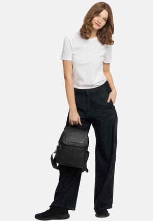 Zaino in pelle nera con finitura testurizzata, dotato di chiusura a patta, tracolla regolabile e dettagli metallici. Il modello indossa una t-shirt bianca e pantaloni scuri.